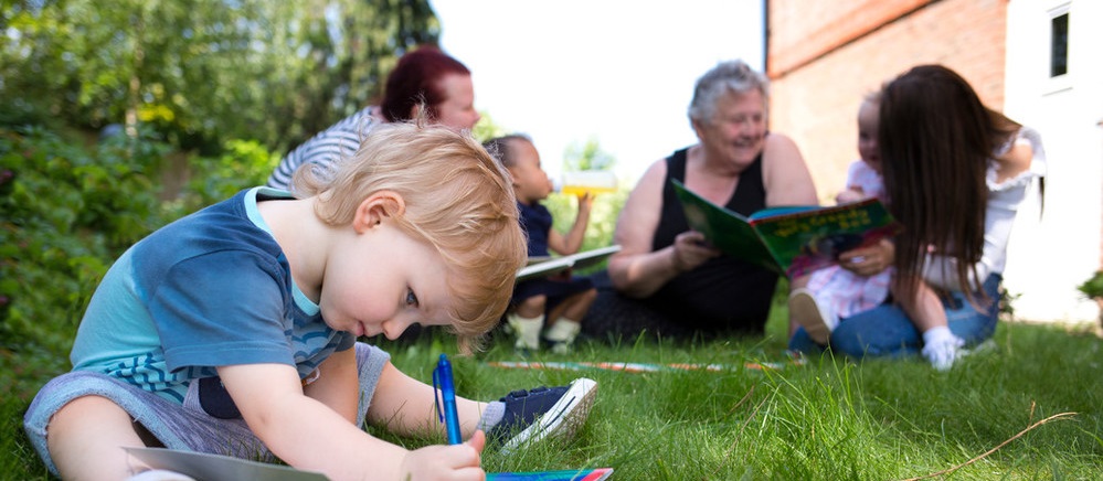 Boy writing on paper in garden with family in background, A2Dominion supported housing services