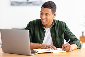 Young man sitting at a laptop in his home making notes
